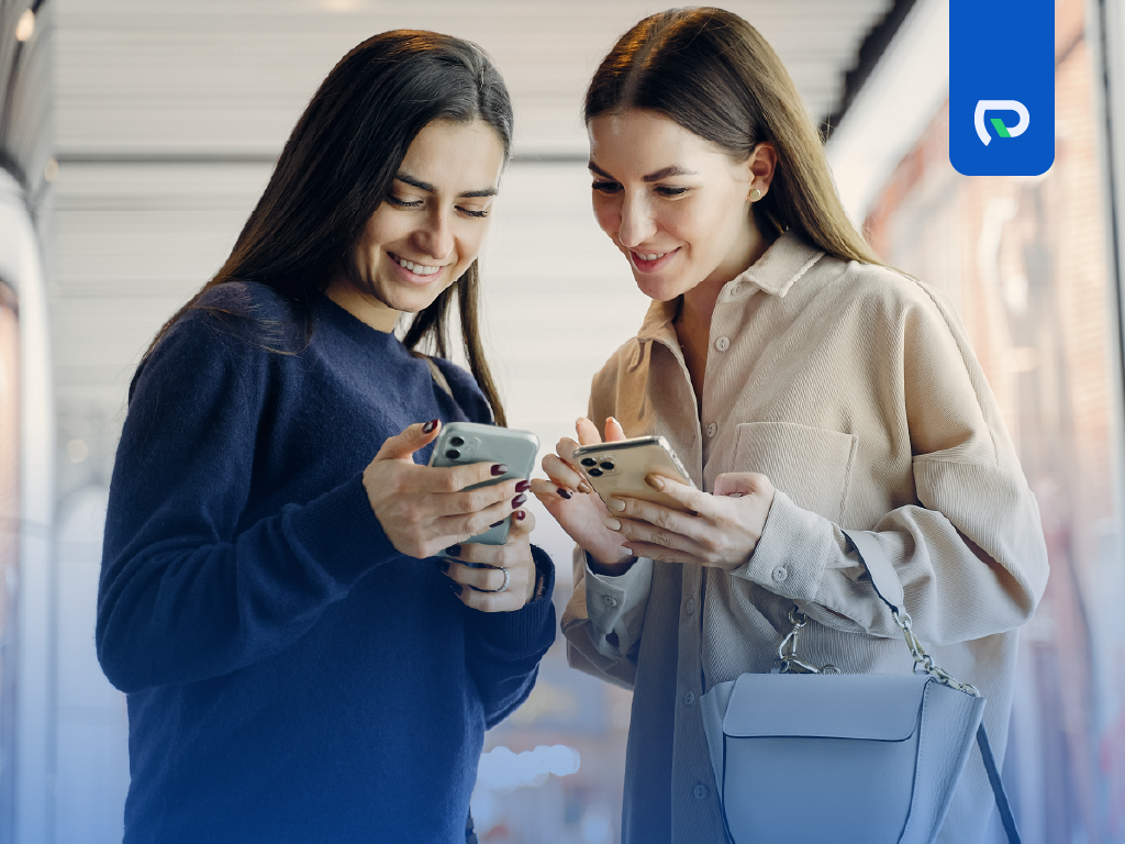 Duas mulheres interagindo com seus smartphones em um ambiente moderno, sorrindo e compartilhando momentos. A imagem destaca a conexão social através da tecnologia.