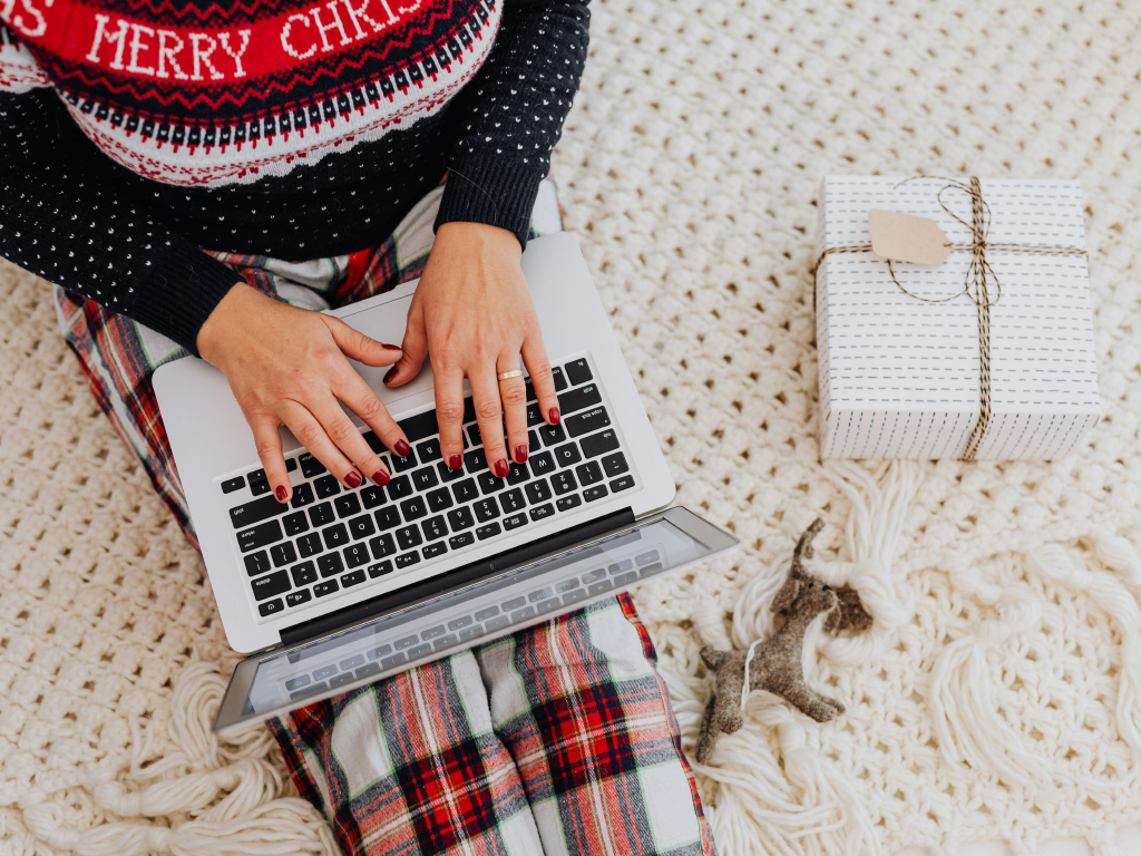 Pessoa usando sweater de Natal e calças xadrez trabalhando em laptop com embalagem de presente ao lado, em ambiente aconchegante de Natal.