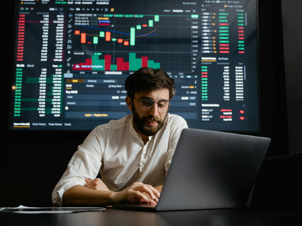 Homem analisando gráficos de ações no computador com tela de mercado financeiro ao fundo, representando negociações e investimentos em bolsa de valores, buscando evitar a queda nas receitas.