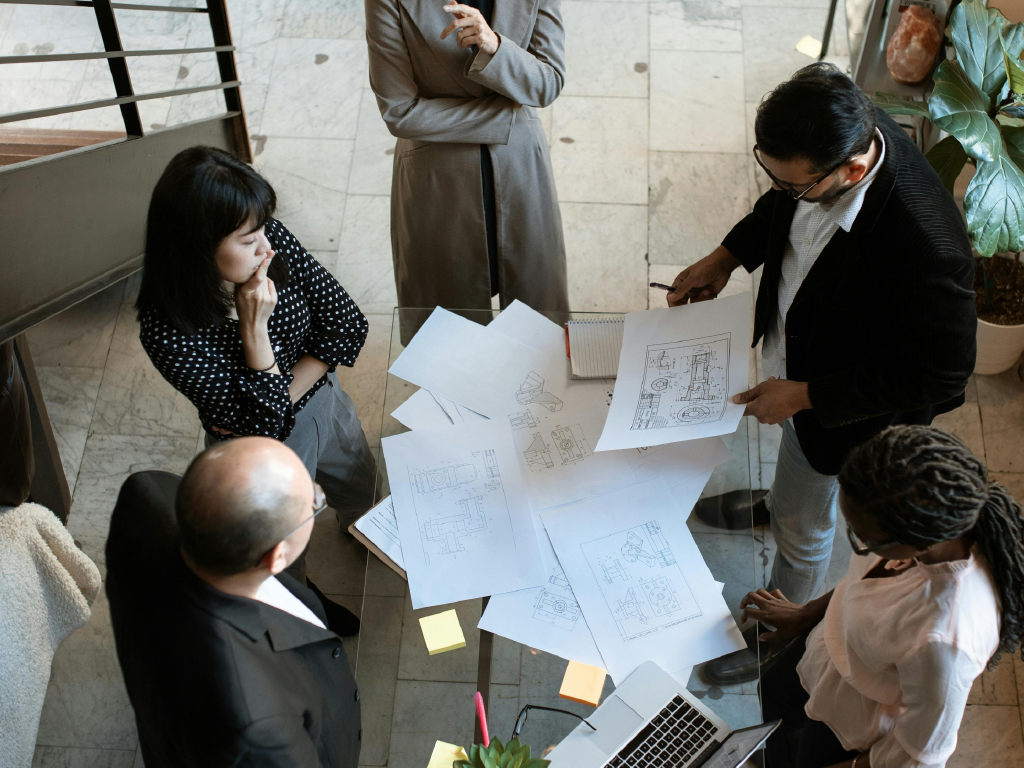 Equipe de profissionais de arquitetura discutindo projetos de construção em uma reunião de trabalho, com desenhos técnicos e um laptop sobre a mesa.