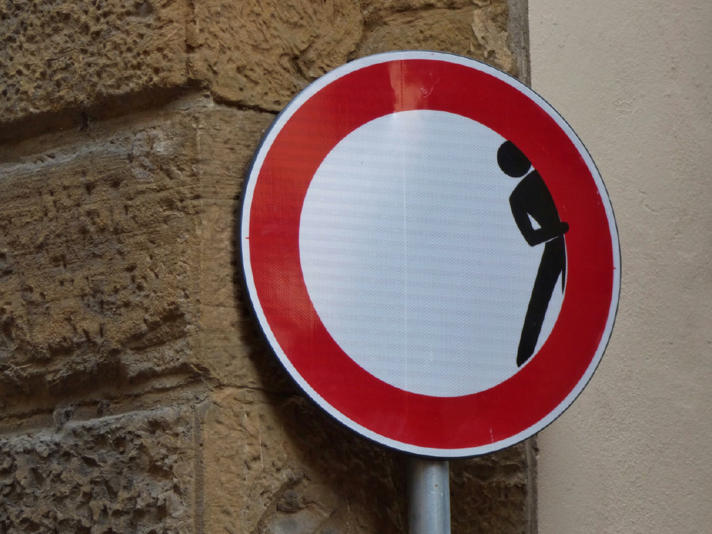 Placa de trânsito humorística com desenho de pessoa se escondendo e símbolo de proibição, localizada em muro de pedra e parede ao fundo, indicando advertência de segurança.