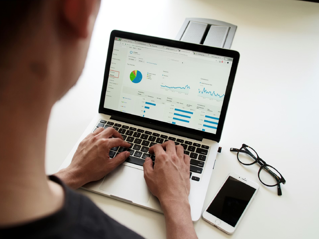 Pessoa analisando gráficos e relatórios de dados em um laptop, com smartphone e óculos ao lado, focada em estratégias de marketing digital.