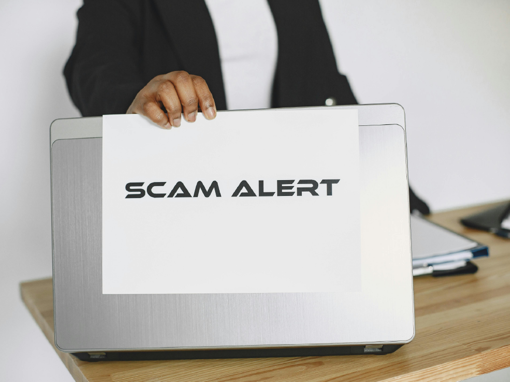 Pessoa segurando um papel com a mensagem 'Scam Alert' em frente a um computador portátil, indicando um alerta de golpe ou fraude online.