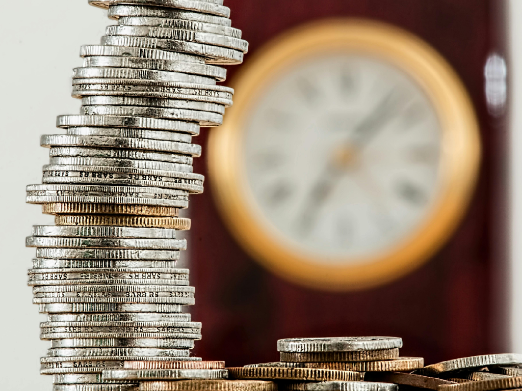 Imagem de uma pilha de moedas variadas ao lado de um relógio de parede amarelo em um fundo desfocado.