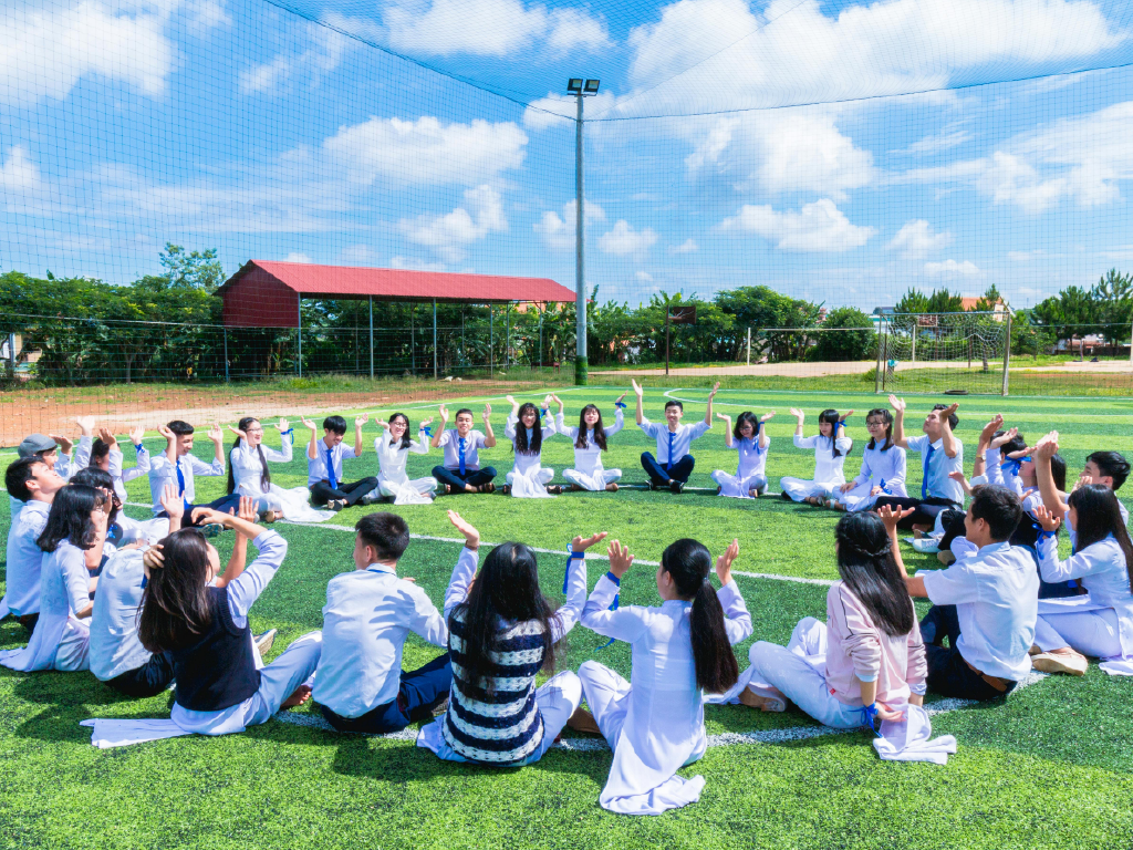 Grupo de estudantes participando de atividade ao ar livre na escola, sentados em círculo na grama, com céu azul e estrutura escolar ao fundo, promovendo aprendizado e convivência.