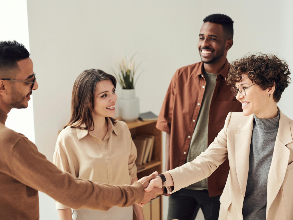 Grupo de pessoas sorridentes em ambiente de escritório, cumprimentando-se com aperto de mãos e demonstrando cordialidade.