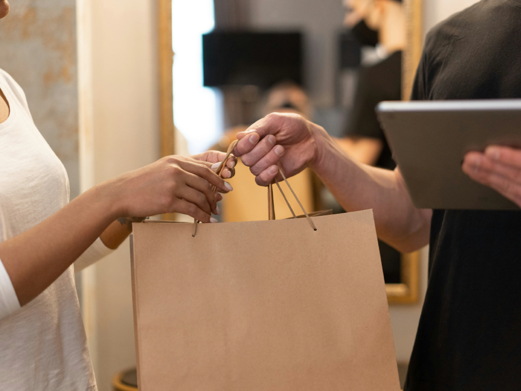 Pessoa recebendo uma sacola de papel de um atendente com tablet, em um ambiente de loja ou comércio, com foco na troca de compras.
