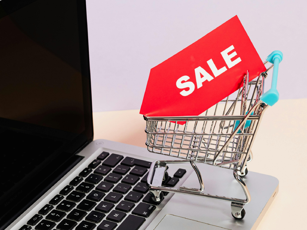 Carrinho de supermercado com etiqueta 'Sale' sobre um teclado de laptop, simbolizando promoções e ofertas online.