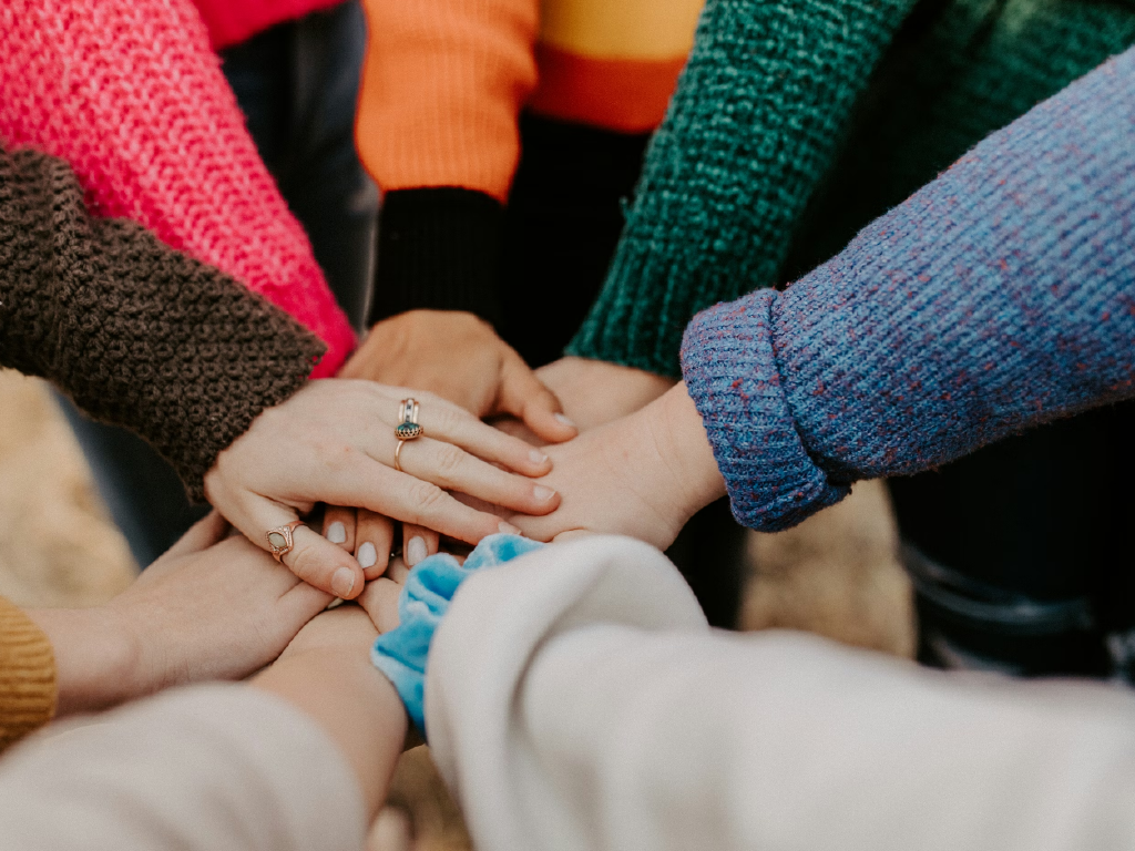 Imagem de várias mãos de pessoas diferentes unidas em um gesto de solidariedade e união, usando roupas coloridas. Representa amizade, apoio e diversidade.