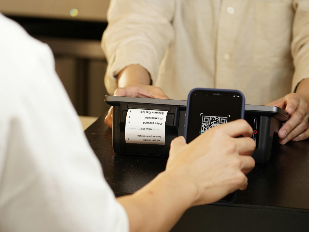 Pessoa usando smartphone para digitalizar um código QR na impressora de recibos, enquanto outro indivíduo observa, em ambiente de caixa ou loja.