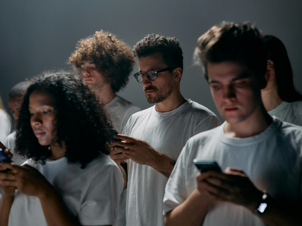 Grupo de jovens usando camisetas brancas, concentrados, todos mexendo em seus smartphones em ambiente com iluminação suave e fundo escuro, representando o trabalho na era digital.