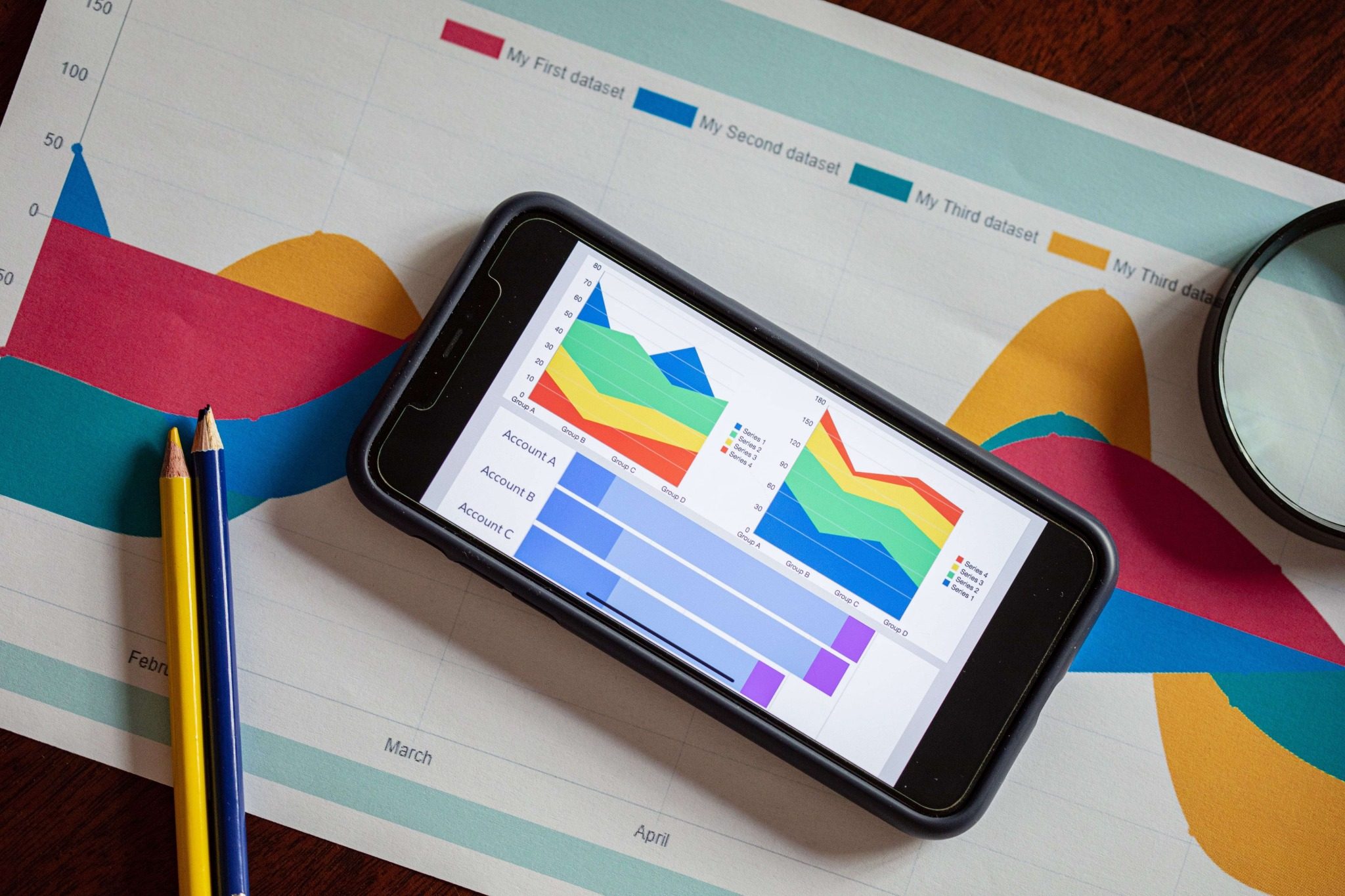 Um smartphone exibindo gráficos de dados coloridos está sobre um gráfico impresso, ao lado de dois lápis e uma lupa em uma superfície de madeira.